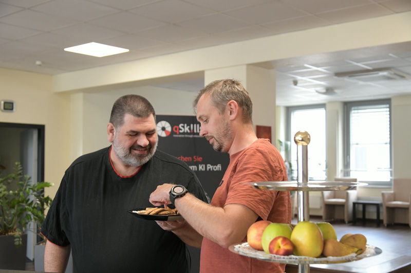 Two male participants in the qSkills Cafe & Lounge area, pausing in front of the fruit basket, eating cookies.