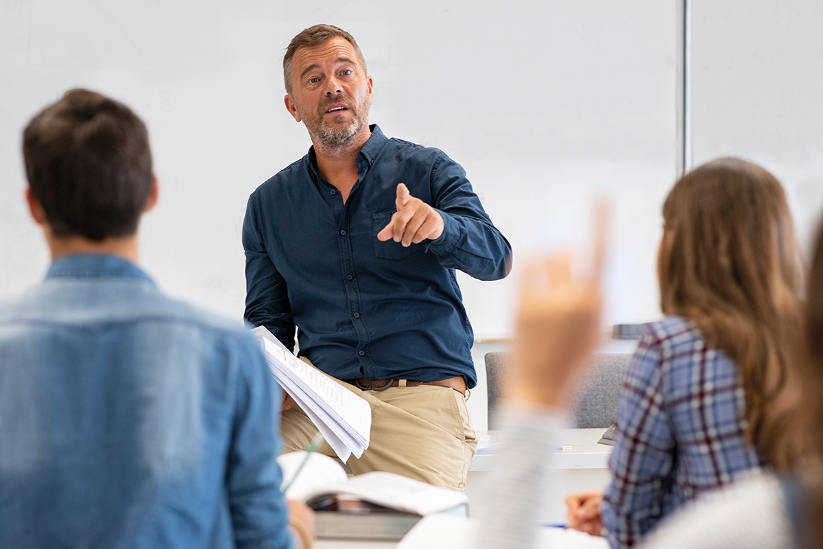 A lecturer with an outstretched index finger speaking to the participants, who are seen from behind.