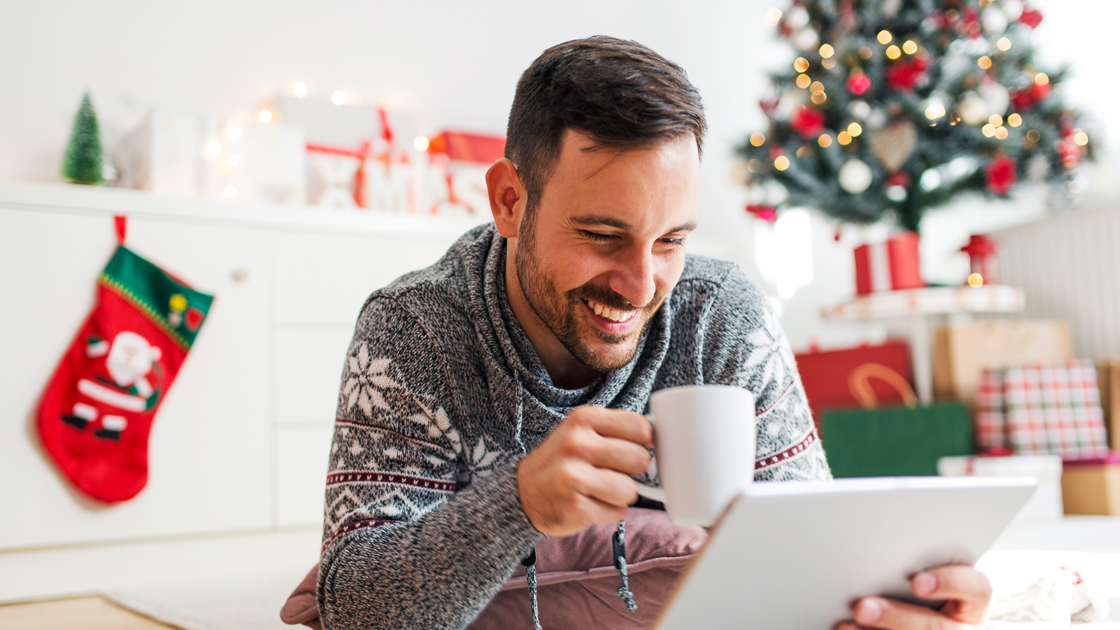 Mann mit Tablet, Tasse - im Hintergrund Weihnachtsbaum und Geschenke