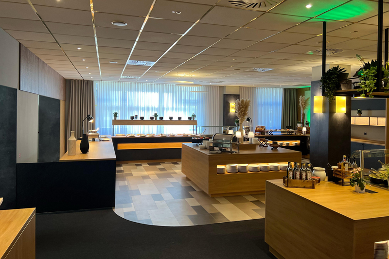 Restaurant area at the NOVINA Hotel am Südwestpark in Nuremberg with salad buffet, food serving area, and dessert area without guests and green ceiling lighting.