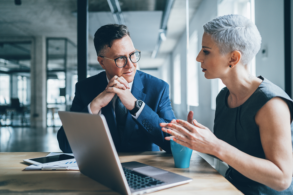 Two businesspeople, a man and a woman, dressed in business attire, sitting in front of an open laptop in a consultation meeting, their hands in front of their bodies, their heads turned toward each other.
