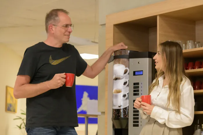 Zwei Teilnehmende, Mann und Frau, am qSkills Kaffeeautomat im Cafe & Loungebereich, die Gesichter zugewandt, im Gespräch bei einer Tasse Kaffee.