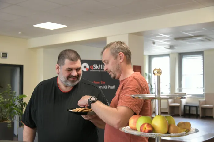 Zwei männliche Teilnehmer im qSkills Cafe & Loungebereich, vorm Obstkorb pausierend, beim Kekse essen.