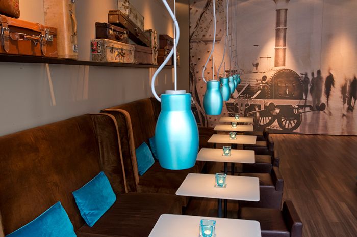 Cozy seating and dining area with benches, chairs, tables, and blue lamps, without guests. In the background, a picture with a train theme in the hotel “Motel One Nuremberg-City.”