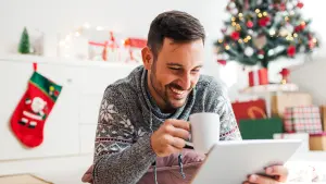 Mann mit Tablet, Tasse - im Hintergrund Weihnachtsbaum und Geschenke