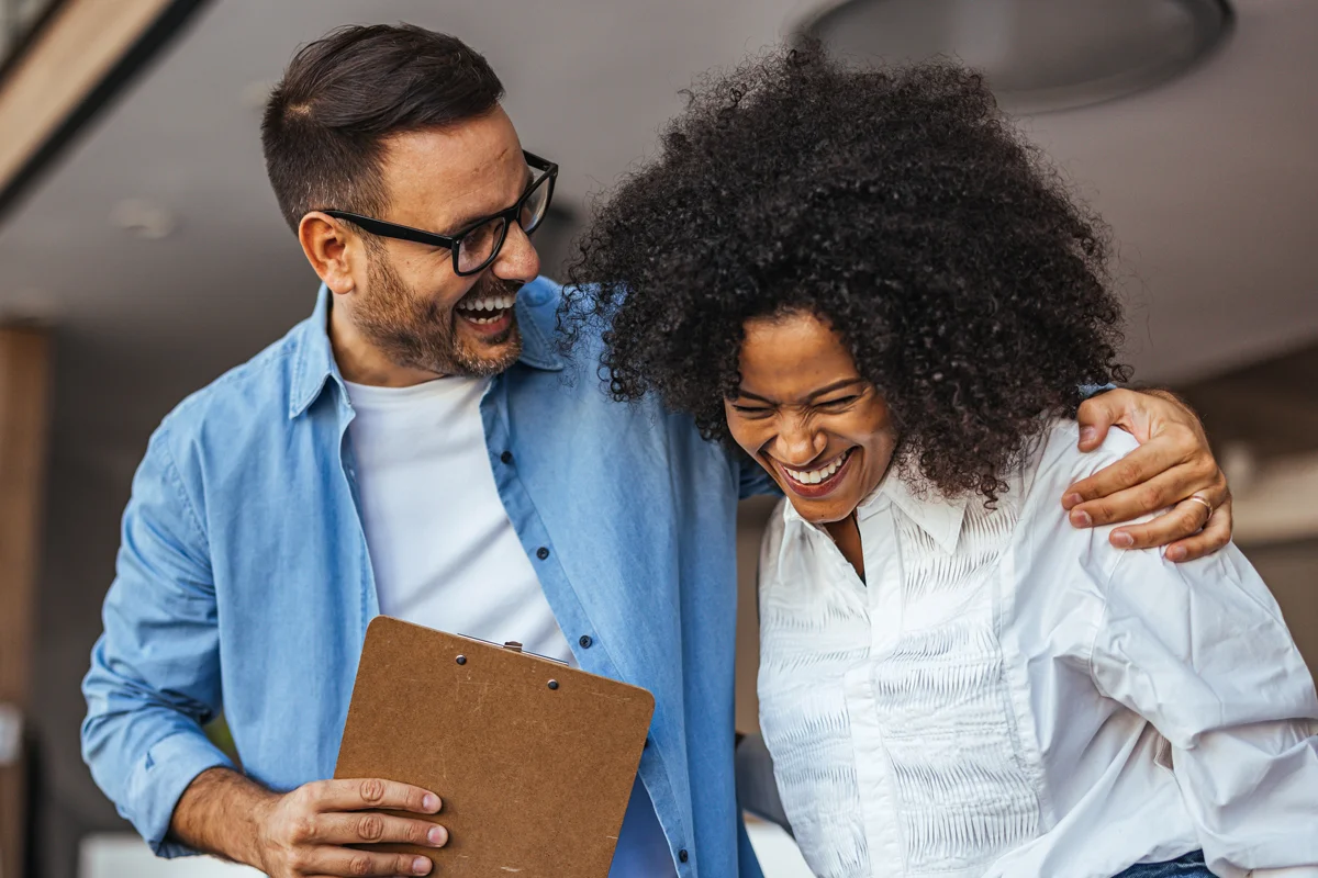 Two colleagues share a joyful moment in a modern office, laughing and enjoying each other's company.