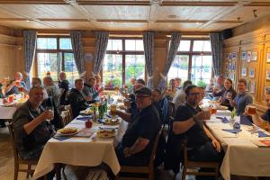 Eine große Gruppe Teilnehmende beim traditionellen qSkills Teilnehmerabend (Montag) im Restaurant "Bratwurst Röslein" beim Abendessen mit gehobenen Gläsern, in ausgelassener Stimmung, sich gegenseitig zuprostend mit Bier.